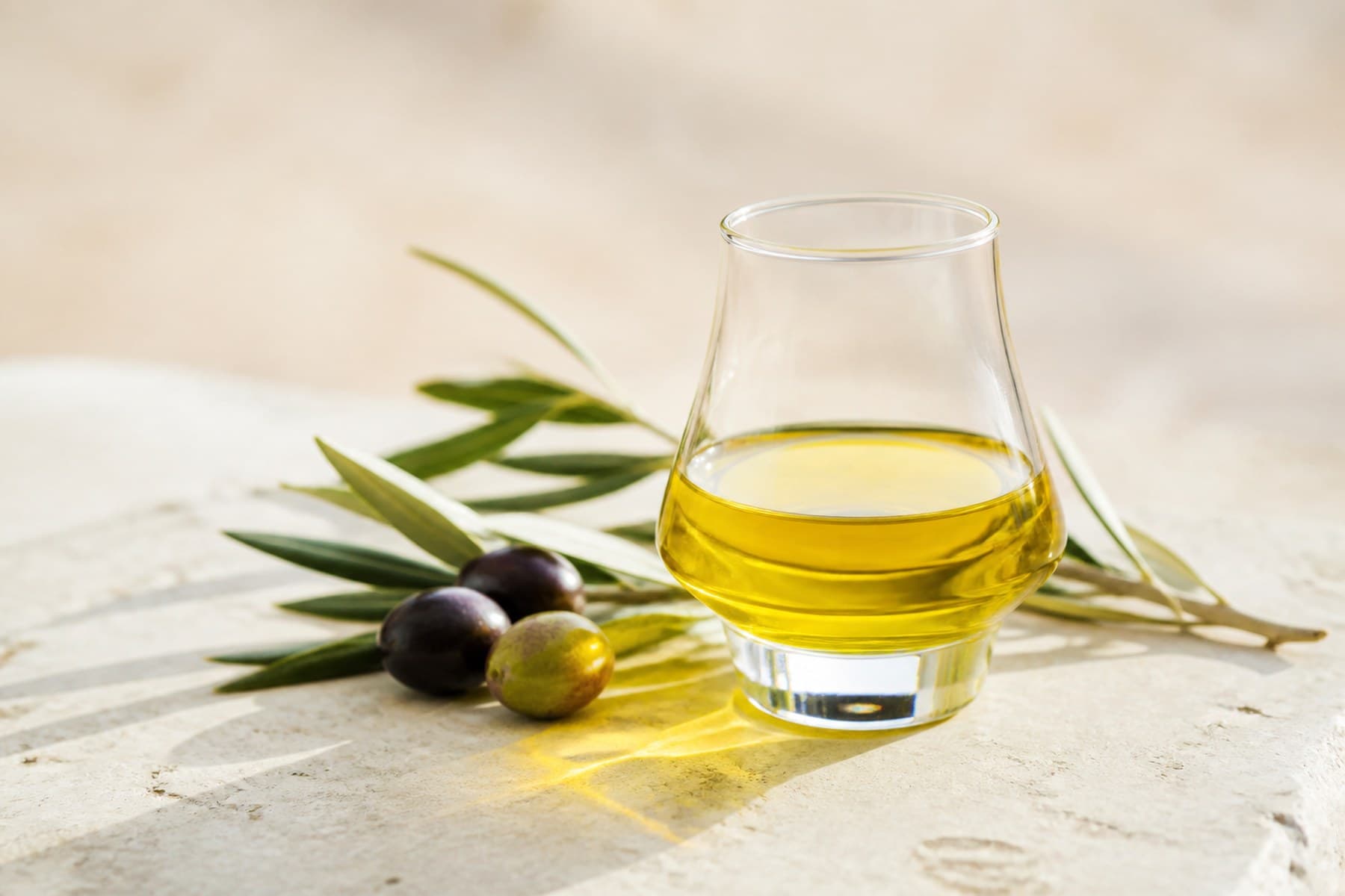 Green-gold extra virgin olive oil in a small clear tasting glass beside olives