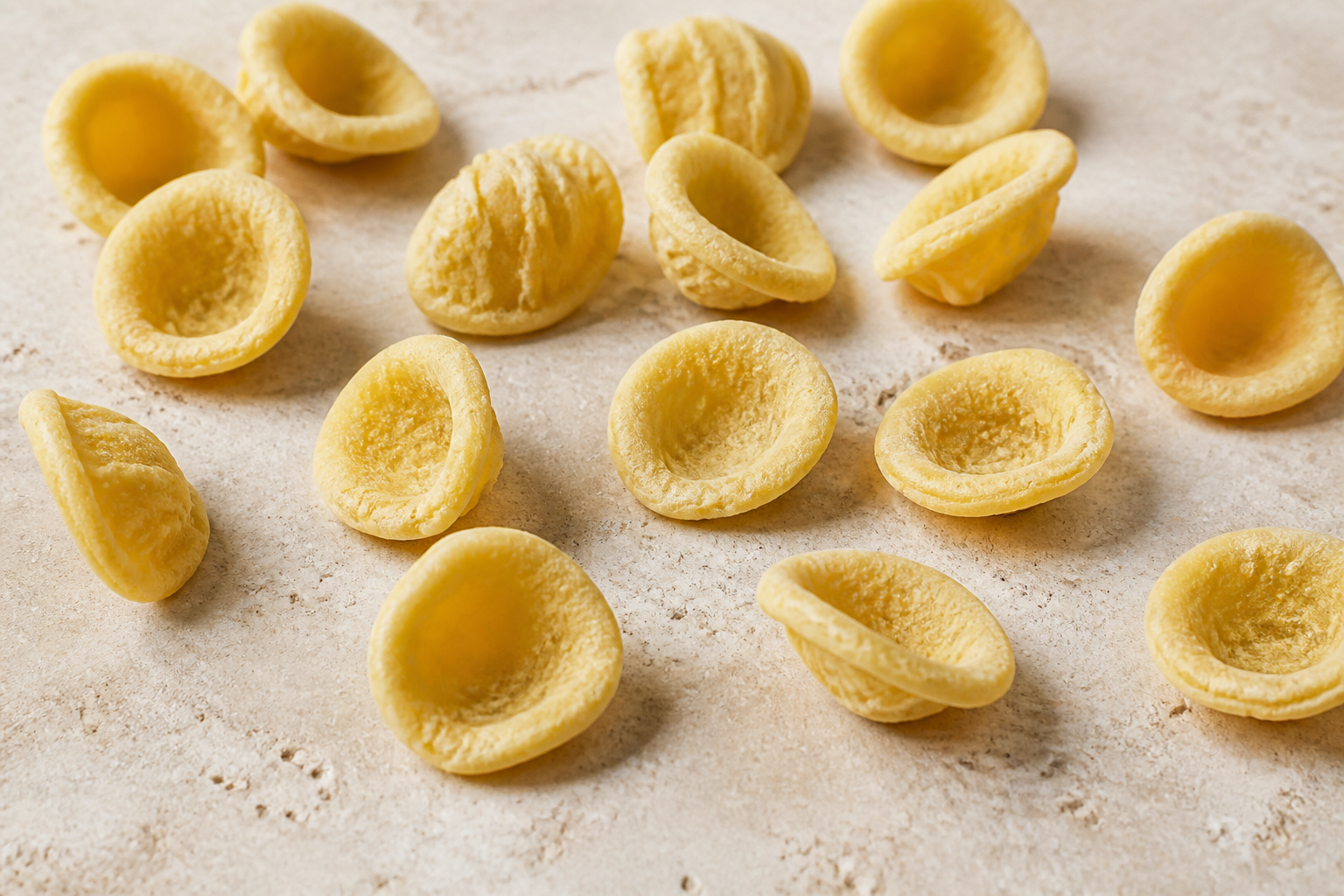 Dry orecchiette pasta showing small rough ear-shaped cups and thick rims on pale stone
