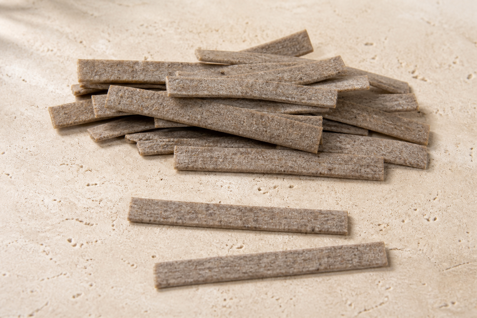 Dry buckwheat pizzoccheri showing short flat grey-brown speckled ribbons on pale stone